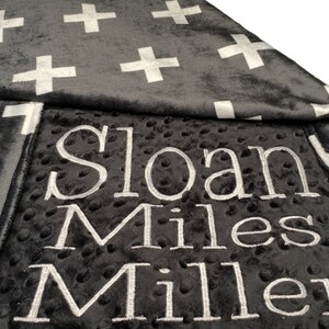 May include: A black and white baby blanket with a white cross pattern on one side and the name "Sloan Miles Miller" embroidered on the other side.