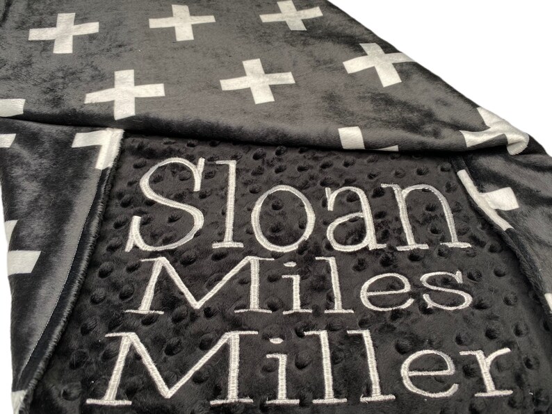 May include: A black and white baby blanket with a white cross pattern on one side and the name "Sloan Miles Miller" embroidered on the other side.