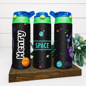 Op de afbeelding: Drie cilindervormige drinkflessen met een zwarte achtergrond in ruimtethema, voorzien van kleurrijke planeten, sterren en sterrenstelsels. De flessen hebben felgroene en blauwe deksels. Eén fles toont de naam Henry, een andere het woord SPACE.
