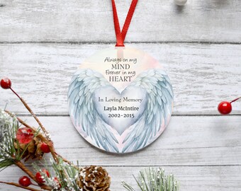 Op de afbeelding: Een witte ronde ornament met een aquarelontwerp van engelenvleugels en een hart. De tekst luidt "Always on my MIND forever in my HEART In Loving Memory Layla McIntire 2002-2015".