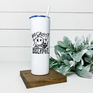 Peut inclure: Un gobelet en acier inoxydable blanc avec un couvercle bleu et un graphique noir qui dit "Not Bossy Just Aggressively Helpful" avec un visage souriant avec des X pour les yeux et une main de squelette faisant le signe du rock and roll.