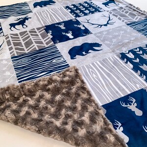 Peut inclure: Une couverture bébé douce, grise et bleue avec un motif patchwork représentant des animaux de la forêt, des flèches et des motifs de grain de bois.