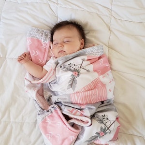 May include: A baby girl sleeping peacefully in a pink and grey patterned blanket featuring arrows and flowers. The blanket is soft and comfortable, ideal for a little one.