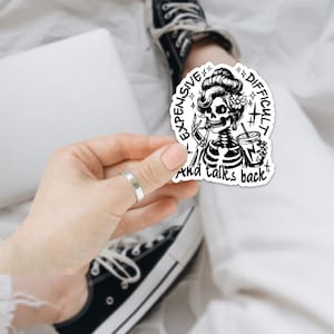 May include: A black and white sticker depicting a skeleton with a vintage hairstyle, holding a coffee cup and a peace sign. The sticker reads "Expensive, Difficult, and talks back" in a circular arrangement.