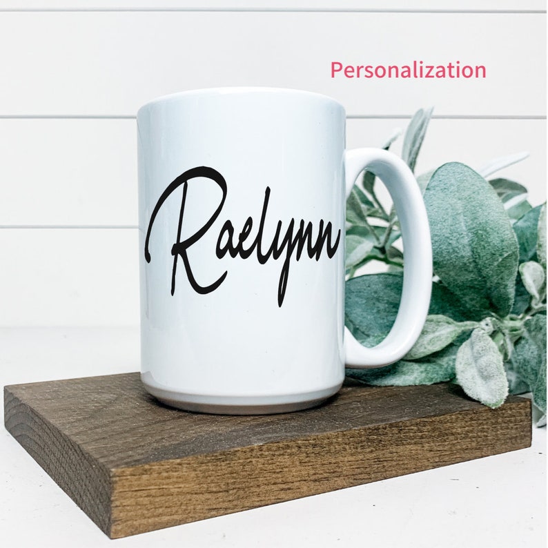 K&ouml;nnte beinhalten: Eine wei&szlig;e Keramiktasse mit dem Namen "Raelynn" in schwarzer Kursivschrift auf der Vorderseite. Die Tasse steht auf einem Untersetzer aus Holz.