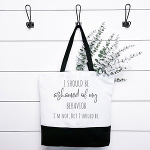 May include: A white canvas tote bag with black handles and a black bottom. The bag has the text "I SHOULD BE ashamed of my BEHAVIOR I'M NOT, BUT I SHOULD BE." printed on the front in black.