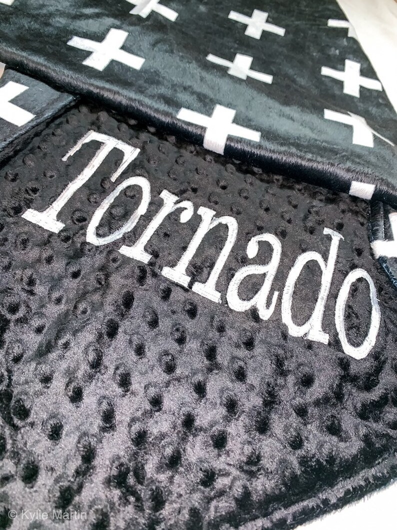 May include: Black and white baby blanket with a white cross pattern on one side and a black minky fabric with the name "Tornado" embroidered in white on the other side.