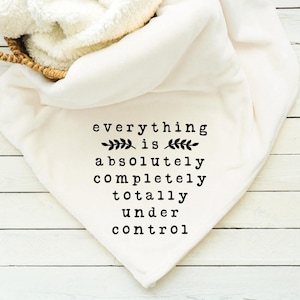 Op de afbeelding: Een witte deken met de tekst "everything is absolutely completely totally under control" in zwarte letters. De deken is opgevouwen en over een houten oppervlak gedrapeerd. Een mand met een wit, pluizig materiaal bevindt zich linksboven.