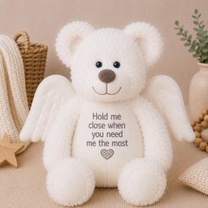 Könnte beinhalten: Ein weißer Plüschteddybär mit weichem, strukturiertem Fell, großen Engelsflügeln auf dem Rücken und einer braunen Nase. Auf der Brust des Bären ist der Text "Hold me close when you need me the most" über einem kleinen Herzsymbol eingestickt.