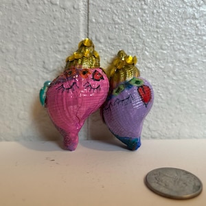 May include: Two hand-painted seashell ornaments. One is pink with a gold top and the other is purple with a gold top. Both have painted faces and are decorated with a heart. A coin is in the foreground.