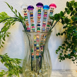 Puede incluir: Un jarrón de cristal transparente contiene seis agitadores de cóctel decorativos. Cada agitador tiene un pomo facetado de diferente color en la parte superior. Los tallos están adornados con cuentas de colores. El follaje verde está dispuesto alrededor del jarrón.