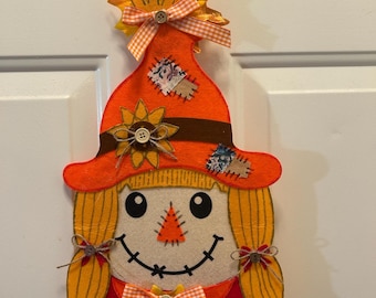 May include: A felt scarecrow decoration with an orange hat and hair, a smiling face, and a red shirt. The hat is adorned with a sunflower, a leaf, and a gingham ribbon. Patches are sewn onto the hat and shirt.