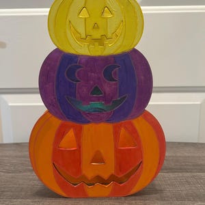 May include: A stacked Halloween decoration featuring three carved pumpkins in yellow, purple, and orange. Each pumpkin has a smiling face with triangular eyes and a carved mouth. The top pumpkin has a green stem.
