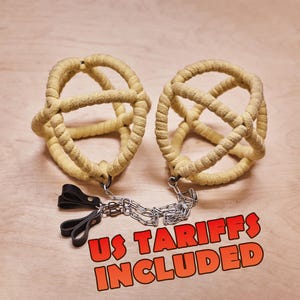 May include: Two spherical restraints constructed from thick, light yellow rope, connected by a silver-toned metal chain with two small black leather loops attached to one end. The text US TARIFFS INCLUDED is printed in orange at the bottom.