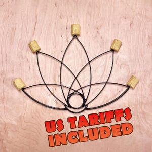 Op de afbeelding: Een zwart metalen vuur-rekwisiet in de vorm van een lotus met vijf beige Kevlar lonten aan de uiteinden van de bloemblaadjes en een centrale ronde handgreep. De tekst US TARIFFS INCLUDED staat in dikke oranje letters onderaan.