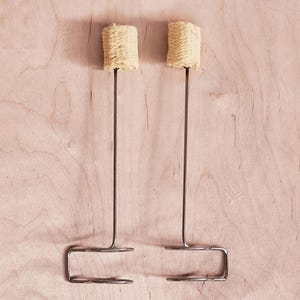 Puede incluir: Dos poi de fuego, un accesorio para bailar con fuego, se muestran sobre un fondo claro de vetas de madera. Cada poi presenta una cabeza cilíndrica tejida unida a una varilla metálica larga y delgada. Las varillas terminan en un asa en forma de U.