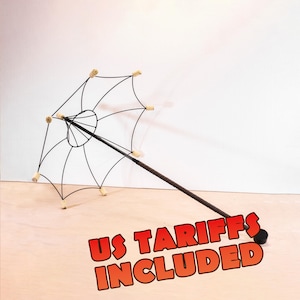 Op de afbeelding: Een paraplu met een zwart draadframe, een lange steel en lichtgekleurde cilindrische uiteinden aan elke spaak. De tekst US TARIFFS INCLUDED staat in grote rode letters met een zwarte omlijning onderaan in de afbeelding weergegeven.