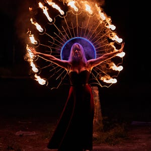 May include: A woman in a red dress performs a fire dance with a circular metal hoop with flames coming from the ends. The hoop is decorated with a blue light.