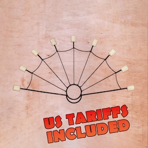 Op de afbeelding: Een zwarte metalen vuurwaaier met acht spaken die uitstralen vanuit een cirkelvormig handvat. Elke spaak eindigt in een cilindrische gebroken witte lont. De tekst US TARIFFS INCLUDED staat in dikke rode letters met een zwarte omlijning.