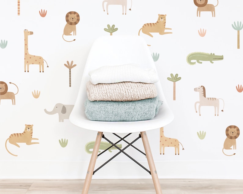 May include: A white molded plastic chair with wooden legs holds a stack of three folded sweaters in white, beige, and light blue. The wall behind features a repeating pattern of illustrated jungle animals including lions, tigers, giraffes, and crocodiles.