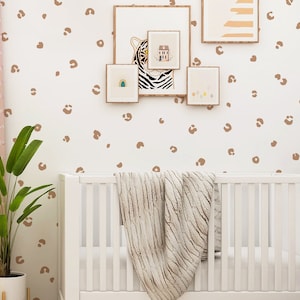 May include: A white crib with a brown and white blanket in a nursery with a white wall featuring brown leopard print decals. There are several framed pictures on the wall.