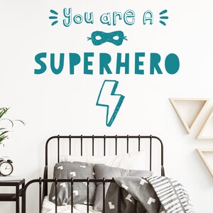 May include: Teal blue wall decal with the text "You are a Superhero" with a mask and lightning bolt graphic. The decal is on a white wall above a black metal bed frame with grey and white bedding.