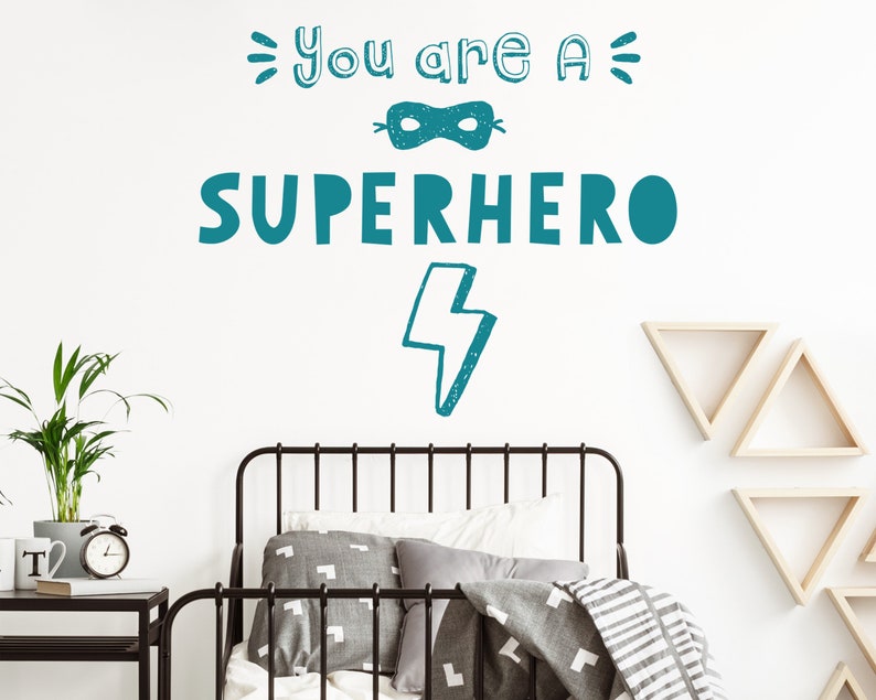 May include: Teal blue wall decal with the text "You are a Superhero" with a mask and lightning bolt graphic. The decal is on a white wall above a black metal bed frame with grey and white bedding.