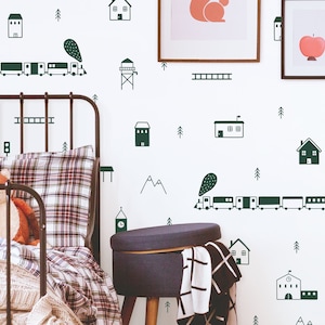 May include: A white wall with green and black illustrations of houses, trees, mountains, a train, and a ladder. A black metal bed frame with a plaid patterned pillow and a white blanket is in the foreground. A dark gray ottoman with a black and white plaid throw is in the lower right corner.