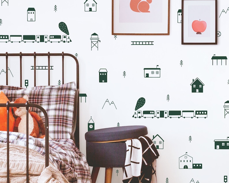 May include: A white wall with green and black illustrations of houses, trees, mountains, a train, and a ladder. A black metal bed frame with a plaid patterned pillow and a white blanket is in the foreground. A dark gray ottoman with a black and white plaid throw is in the lower right corner.