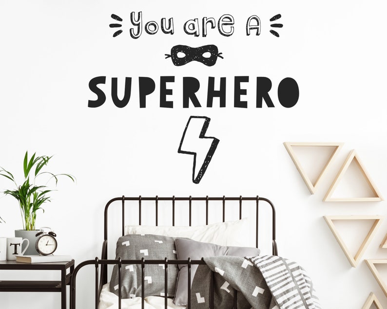 May include: Black and white wall decal with the text "You are a SUPERHERO" and a lightning bolt. The decal is on a white wall above a black metal bed frame with grey bedding.