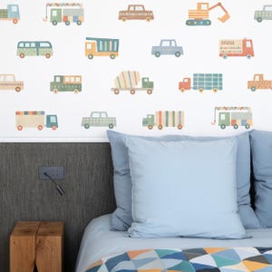 May include: A white wall with colorful vehicle decals, including a truck, a cement mixer, a crane, and a bus. The decals are arranged in a random pattern and are all different sizes. The wall is behind a bed with blue bedding and a blue pillow.