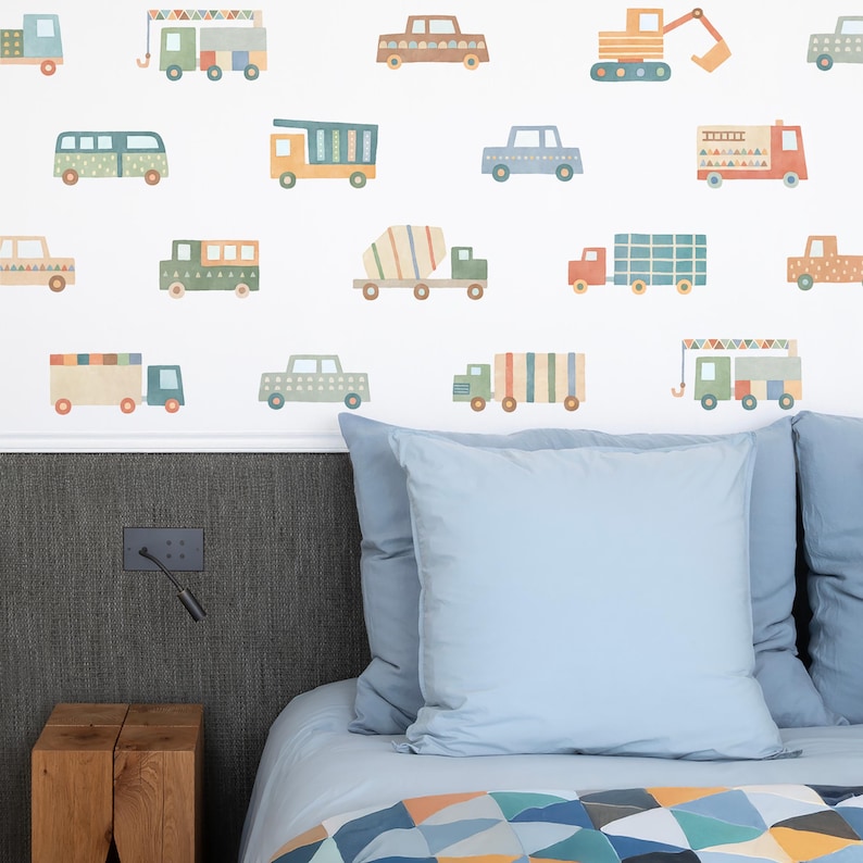 May include: A white wall with colorful vehicle decals, including a truck, a cement mixer, a crane, and a bus. The decals are arranged in a random pattern and are all different sizes. The wall is behind a bed with blue bedding and a blue pillow.