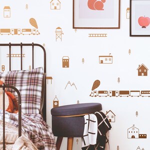 May include: A brown and white train with a brown and white smoke plume travels across a white wall with brown house and tree illustrations. A black metal bed frame with a plaid comforter and a blue ottoman are in the foreground.
