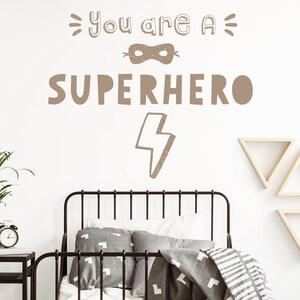 May include: A brown wall decal with the text "You are a Superhero" featuring a mask and lightning bolt graphic. The decal is on a white wall in a bedroom with a bed, bedside table, and plant.