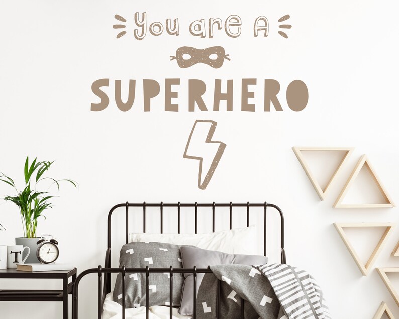May include: A brown wall decal with the text "You are a Superhero" featuring a mask and lightning bolt graphic. The decal is on a white wall in a bedroom with a bed, bedside table, and plant.