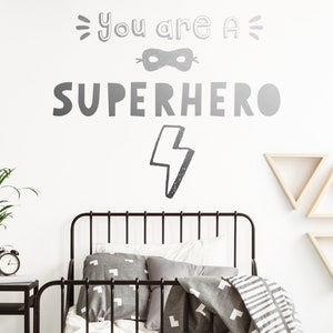 May include: A white wall with a black and white graphic that says "You are a Superhero" with a lightning bolt and a mask. A black metal bed frame with grey bedding and pillows is in front of the wall. There are three wooden triangle shelves on the wall.