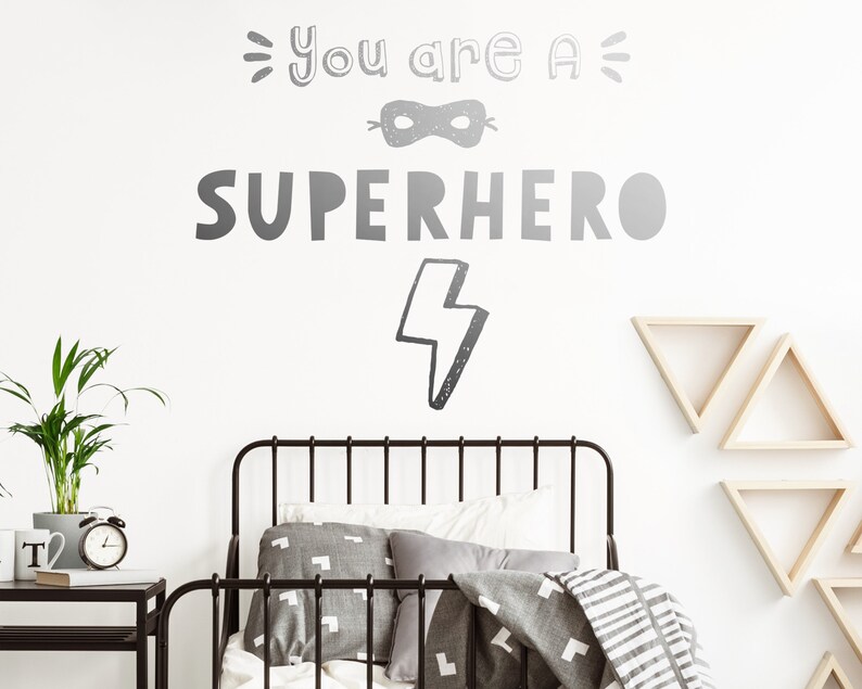May include: A white wall with a black and white graphic that says "You are a Superhero" with a lightning bolt and a mask. A black metal bed frame with grey bedding and pillows is in front of the wall. There are three wooden triangle shelves on the wall.