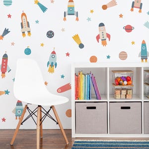Könnte beinhalten: Ein Wandaufkleber-Set mit bunten Illustrationen zum Thema Weltraum, darunter Raketen, Planeten und Sterne in Blau-, Orange- und Rottönen. Die Aufkleber sind auf einer weißen Wand hinter einem weißen Stuhl und einem weißen Regal angebracht.