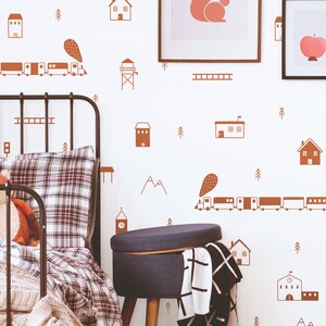 May include: A white wall with brown train, house, and tree decals. A black metal bed frame with a plaid comforter and a blue ottoman are in the foreground.