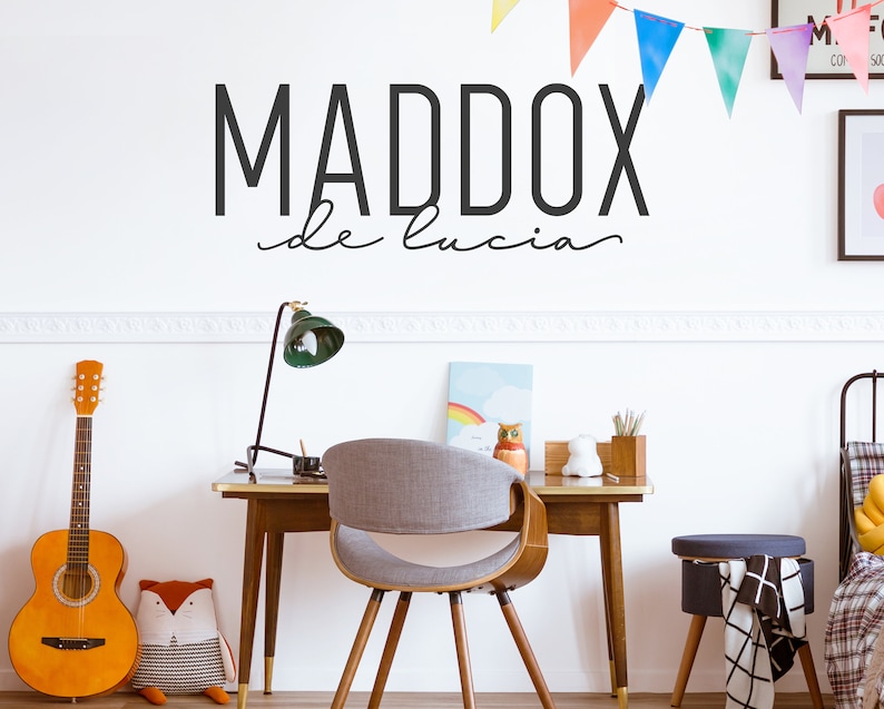 Puede incluir: Una pared blanca con un letrero de nombre en blanco y negro que dice "MADDOX de lucia". La pared est&aacute; decorada con una hilera de banderas de colores. Un escritorio de madera con una silla y una l&aacute;mpara est&aacute; en primer plano. Una guitarra y un animal de peluche est&aacute;n en el suelo.