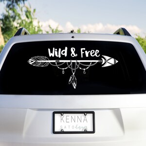 May include: A white car with a black rear window. The window has a white decal with an arrow and the text "Wild & Free". The car has a license plate that reads "KENNA SATODESIGNS".