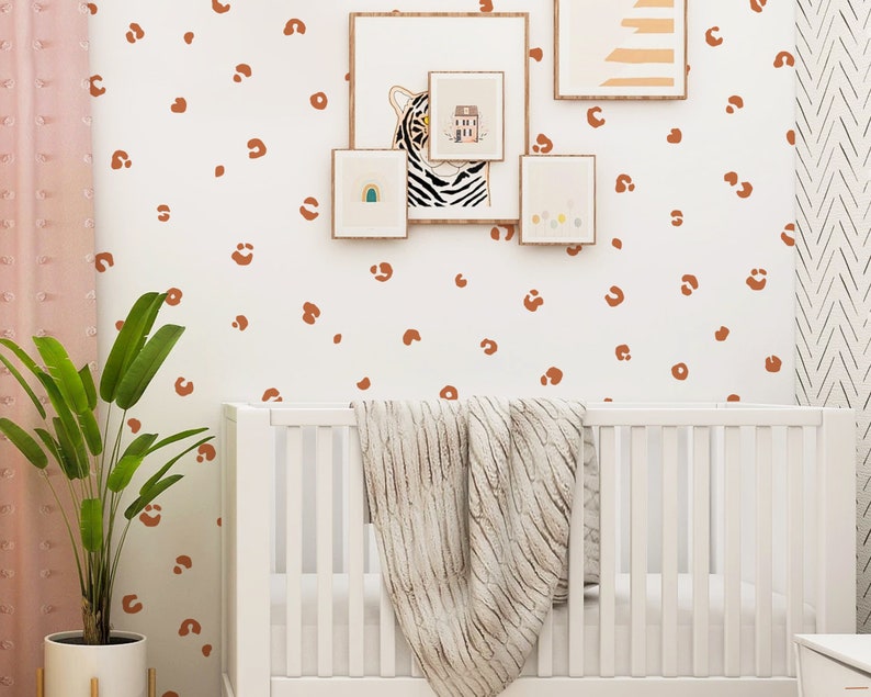 May include: A white crib with a white blanket and a white wall with brown leopard print decals. There are framed pictures on the wall above the crib.