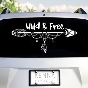 May include: White vinyl decal with an arrow and the words "Wild & Free" on a black background. The arrow has feathers and beads hanging from it. The decal is on the back window of a silver car.