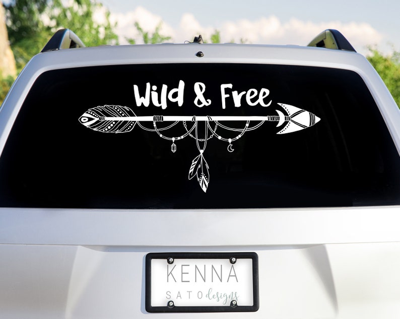 May include: White vinyl decal with an arrow and the words "Wild & Free" on a black background. The arrow has feathers and beads hanging from it. The decal is on the back window of a silver car.