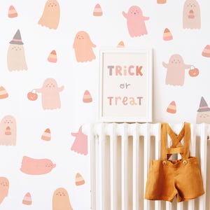 Puede incluir: Vinilos decorativos con fantasmas rosas y color melocot&oacute;n y patrones de ma&iacute;z dulce sobre una pared blanca. Un cuadro con el texto TRICK or TREAT est&aacute; centrado sobre un radiador blanco del que cuelga un peto de lino color mostaza.