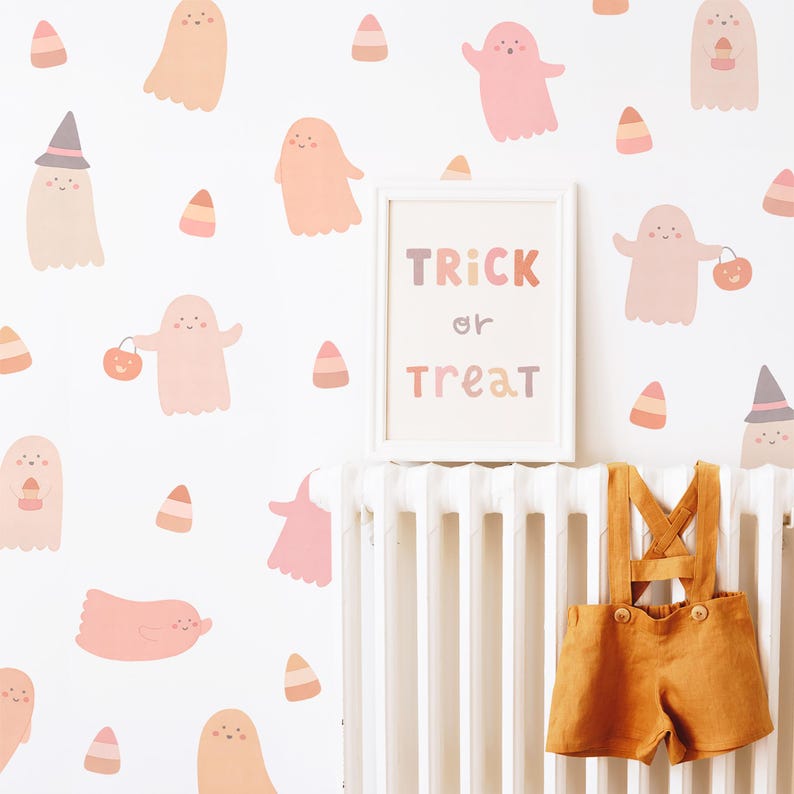 Puede incluir: Vinilos decorativos con fantasmas rosas y color melocot&oacute;n y patrones de ma&iacute;z dulce sobre una pared blanca. Un cuadro con el texto TRICK or TREAT est&aacute; centrado sobre un radiador blanco del que cuelga un peto de lino color mostaza.
