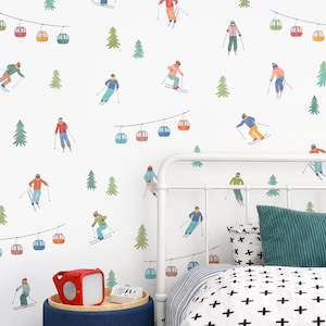 Op de afbeelding: Een muursticker set met een herhalend patroon van skiërs in kleurrijke winterkleding, groene dennenbomen en veelkleurige kabelbaangondels die aan dunne lijnen hangen tegen een witte achtergrond, aangebracht op een slaapkamermuur.