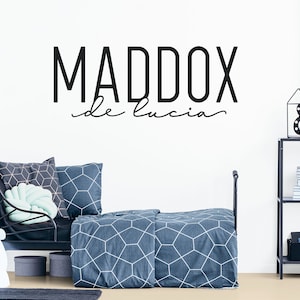 Puede incluir: Pegatina de pared en blanco y negro con el nombre "MADDOX de lucia" en una fuente moderna. La pegatina se muestra en la habitaci&oacute;n de un ni&ntilde;o con una cama de metal negro y ropa de cama con estampado azul y blanco.
