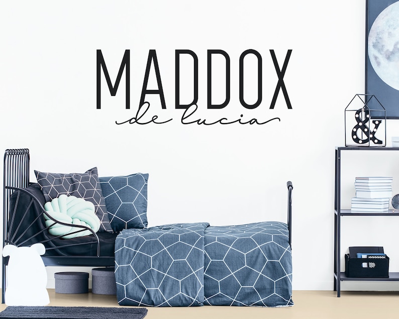 Puede incluir: Pegatina de pared en blanco y negro con el nombre "MADDOX de lucia" en una fuente moderna. La pegatina se muestra en la habitaci&oacute;n de un ni&ntilde;o con una cama de metal negro y ropa de cama con estampado azul y blanco.