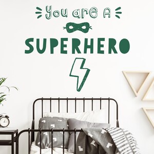 May include: Green and white wall decal with the text "You are a Superhero" and a lightning bolt. The decal is on a white wall above a black metal bed frame with grey bedding.